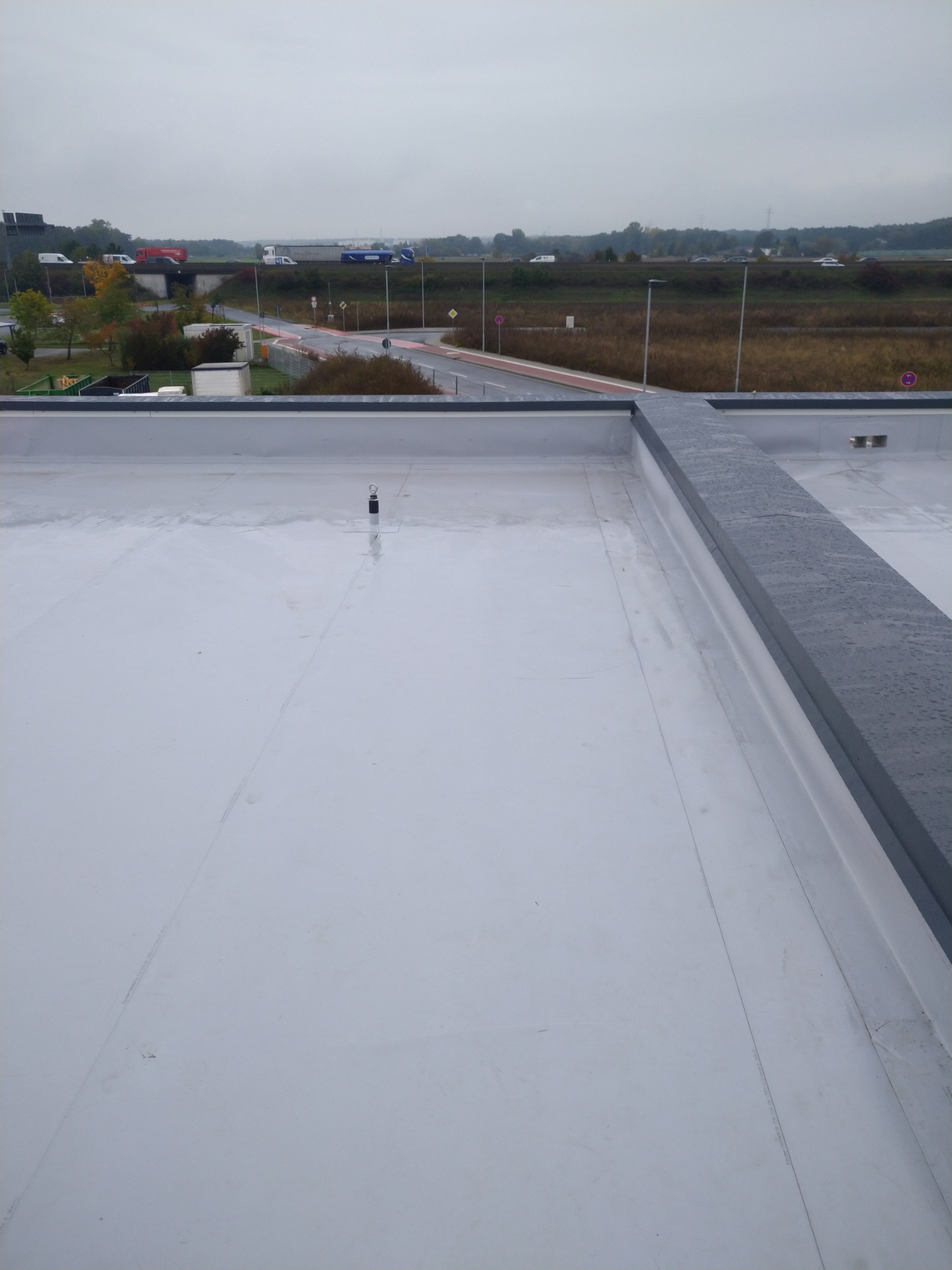 Widok z góry na płaski, biały dach z widoczną szarą attyką i elementami instalacji, w tle krajobraz z drogą i pojazdami w pochmurny dzień.