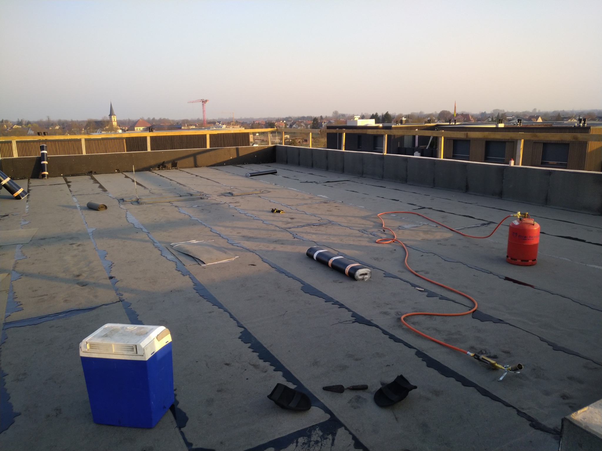 Prace na dachu płaskim: częściowo położona papa, rolka papy, butla gazowa, palnik dekarski, kielnia, niebieska lodówka turystyczna, w tle zabudowa miejska i kościół.