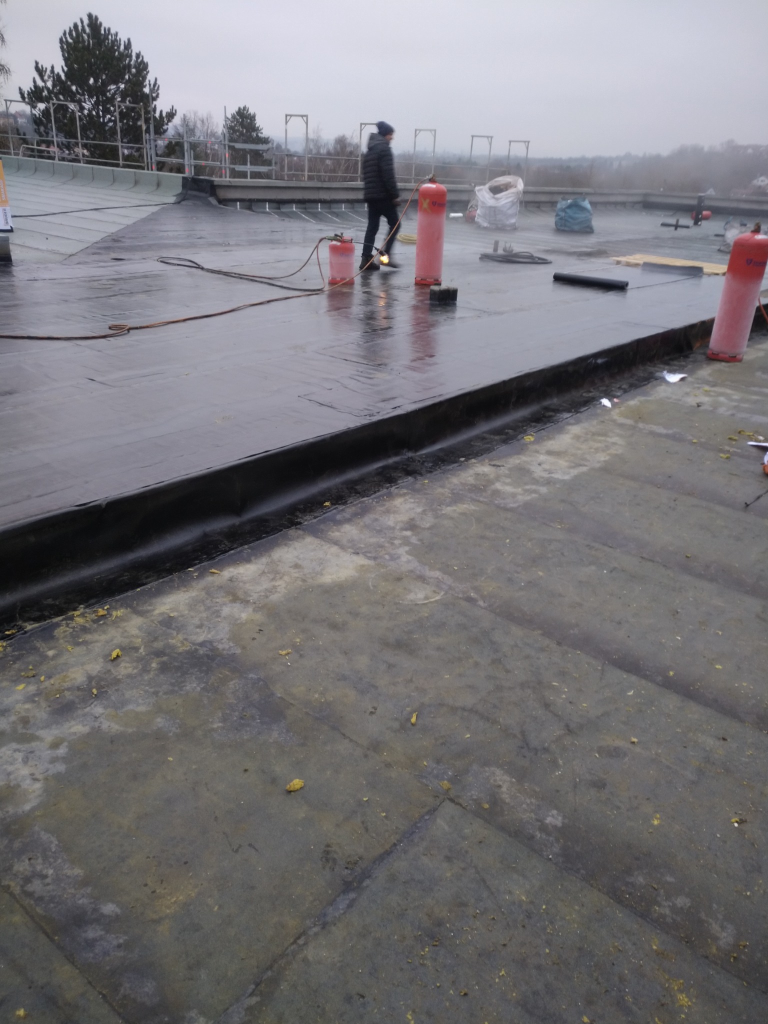 Prace dekarskie na płaskim dachu: dekarz z palnikiem i butlami gazowymi podczas uszczelniania papą bitumiczną na mokrym dachu. Widoczne narzędzia i materiały budowlane.