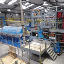 Kompleksowa instalacja przemysłowa z niebieskim zbiornikiem i systemem rur w hali produkcyjnej, widok z góry na platformę roboczą z paletą w tle.