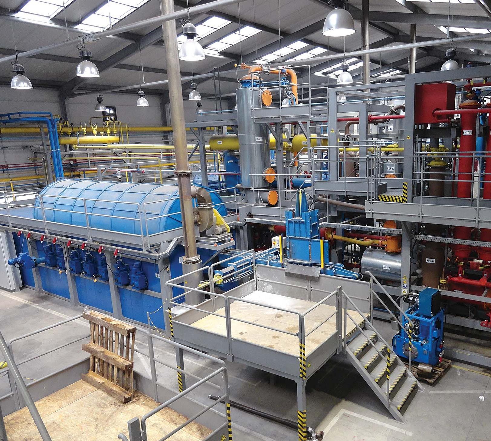 Kompleksowa instalacja przemysłowa z niebieskim zbiornikiem i systemem rur w hali produkcyjnej, widok z góry na platformę roboczą z paletą w tle.