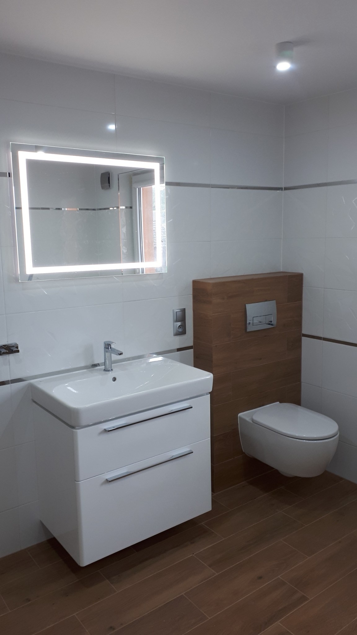 Nowoczesna łazienka z podwieszaną toaletą, szafką umywalkową z szufladami i lustrem z podświetleniem LED, wykończona białymi płytkami z szarym paskiem i drewnopodobną zabudową geberit.