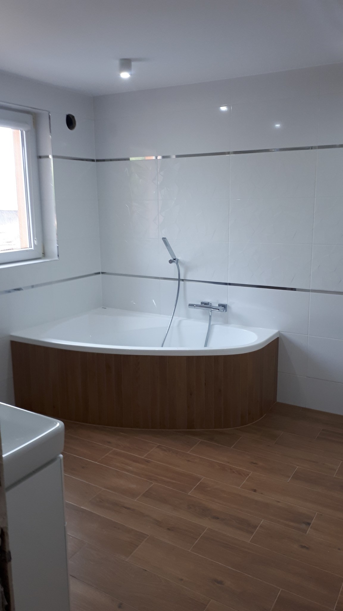 Nowoczesna łazienka z narożną wanną obudowaną panelami imitującymi drewno, białe płytki z metalowymi listwami, brązowa podłoga imitująca drewno, okno po lewej stronie.