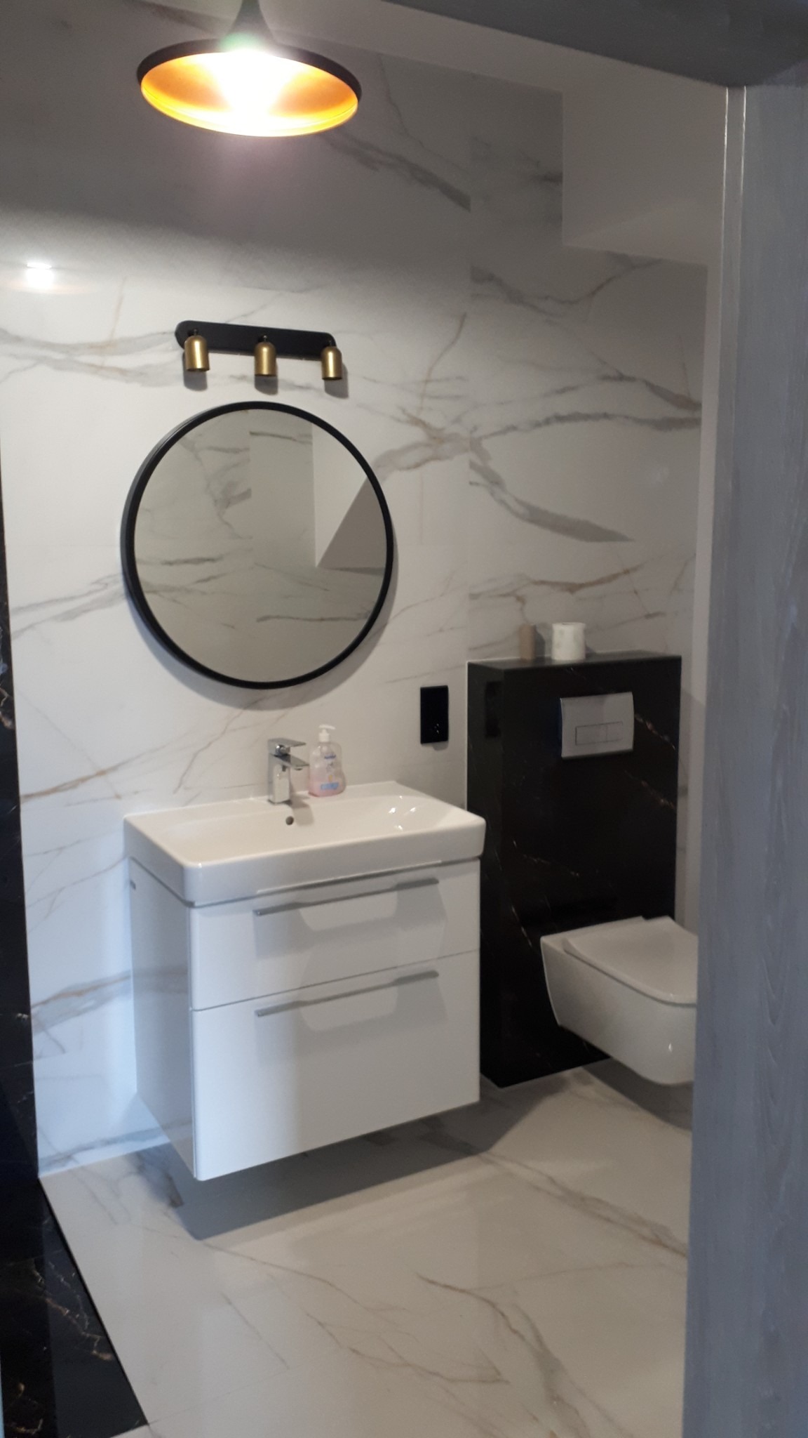Nowoczesna łazienka z białymi płytkami imitującymi marmur, okrągłym lustrem z czarną ramą, białą szafką pod umywalką z szufladami i podwieszanym WC w czarnej obudowie.