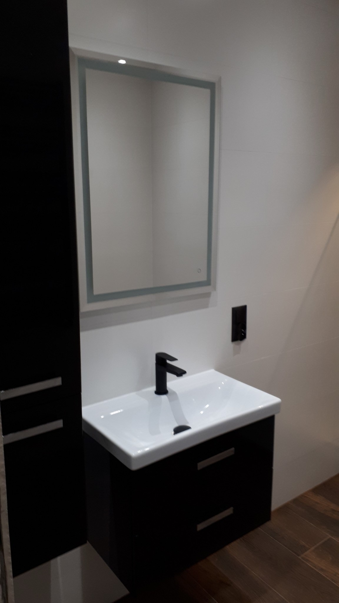 Nowoczesna łazienka z białą umywalką na czarnej szafce z szufladami, czarną armaturą i podświetlanym lustrem na białej ścianie z płytek.