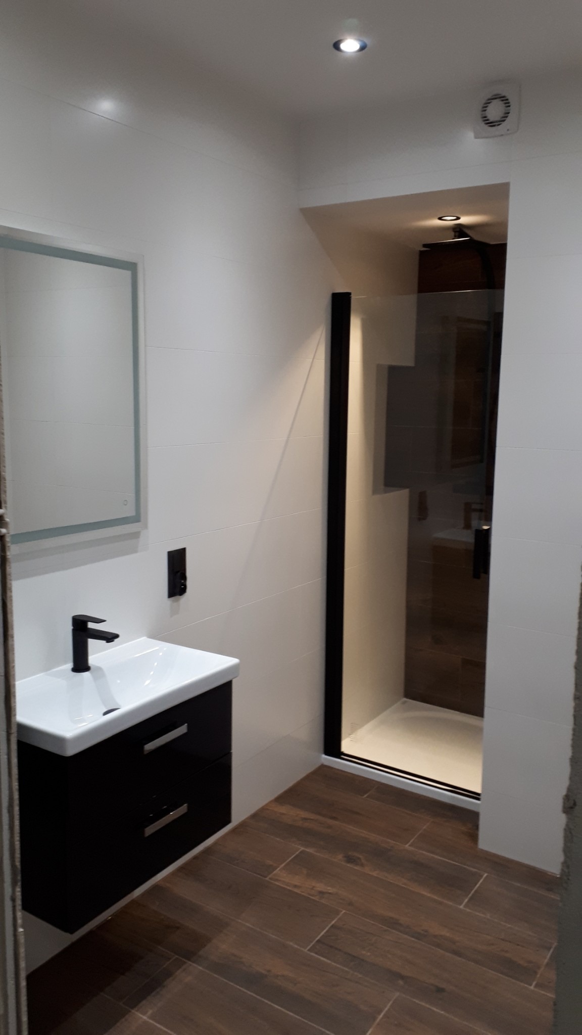 Nowoczesna łazienka z czarną armaturą, kabiną prysznicową typu walk-in z czarną ramą i drewnopodobną podłogą, wykończona w bieli.