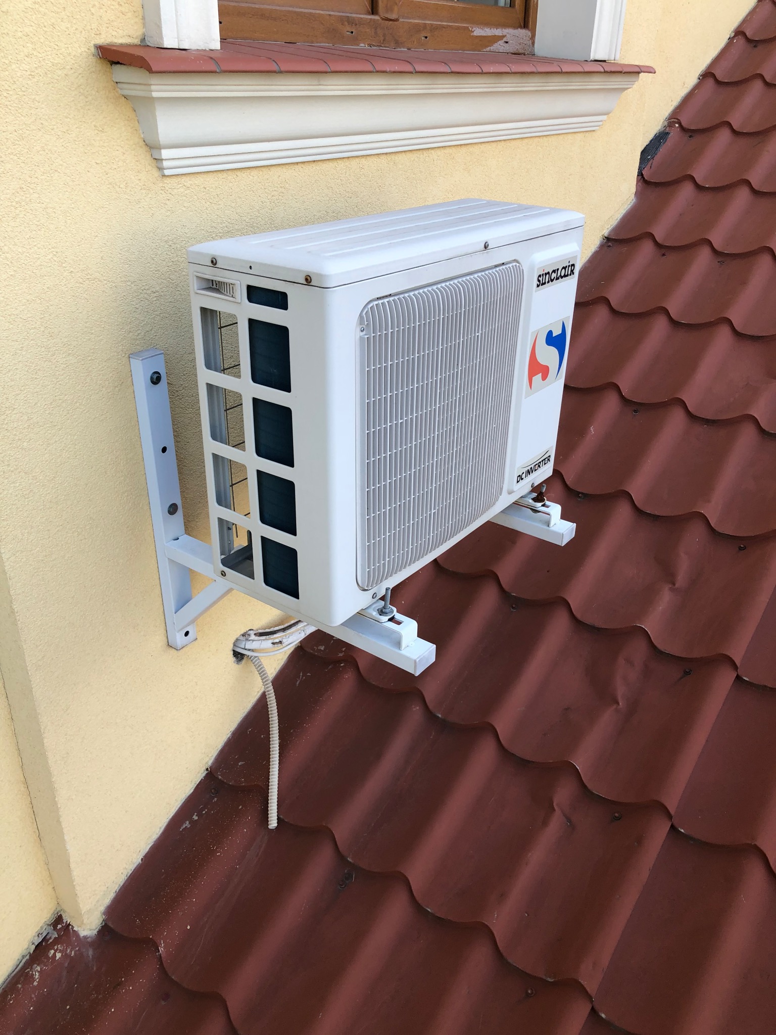 Zewnętrzna jednostka klimatyzacji Sinclair zamontowana na ścianie budynku z żółtą elewacją i czerwoną dachówką. Widoczny przewód elektryczny.