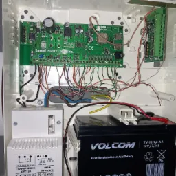 Otwarta obudowa centrali alarmowej Satel Perfecta 16 WRL z widoczną elektroniką, transformatorem Pulsar AWT150 i akumulatorem Volcom 12V/7.2Ah, z podłączonymi przewodami.