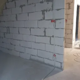 Surowa ściana z betonu komórkowego w trakcie budowy, z widocznymi przewodami elektrycznymi i puszkami instalacyjnymi, surowy beton na podłodze, widok na kolejne pomieszczenie w tle.