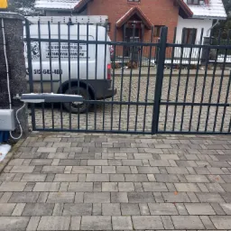 Automatyczna brama przesuwna z napędem, widoczny pojazd serwisowy z logo firmy instalacyjnej w tle, kostka brukowa na podjeździe, częściowo pokryta śniegiem.