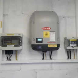 Szary inwerter Fronius SYMO z wyświetlaczem i naklejkami ostrzegawczymi zamontowany na białej, ceglanej ścianie, obok znajduje się skrzynka elektryczna z widocznymi bezpiecznikami i przewodami.