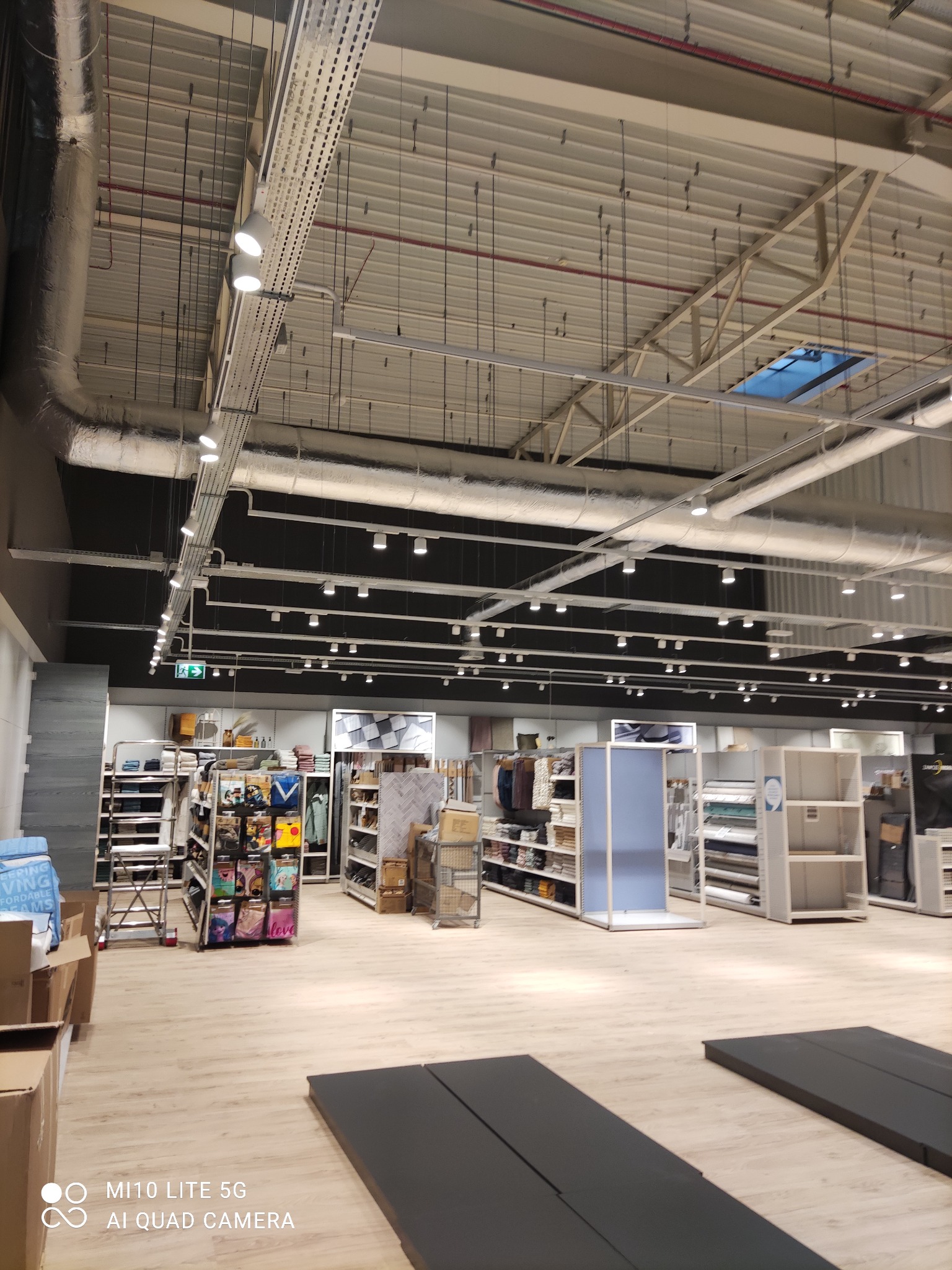 Wnętrze sklepu z widocznymi instalacjami pod sufitem: rury wentylacyjne, korytka kablowe z oświetleniem punktowym, kratownica stropowa i świetliki.