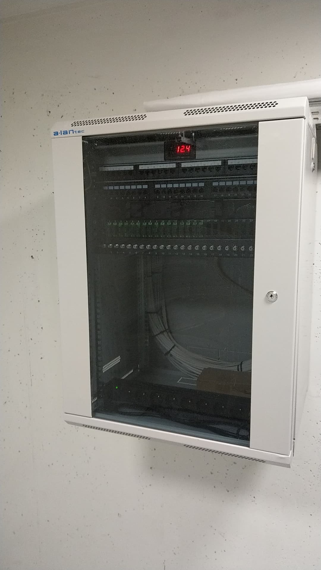 Biała szafa rackowa firmy a.lantec z zamontowanymi patch panelami i uporządkowanymi kablami, wyświetlacz LED pokazuje temperaturę 12.4 stopnie Celsjusza, całość zamontowana na szarej ścianie.