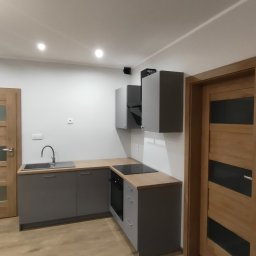 Usług remotowo-wykończeniowe MAG - Nowoczesna, szara zabudowa kuchenna z drewnianym blatem i płytą indukcyjną, widok na drewnianą podłogę i drzwi z przeszkleniem, oświetlenie punktowe sufitowe.