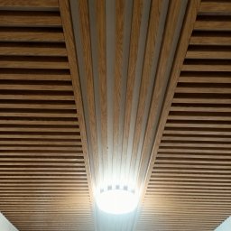 Usług remotowo-wykończeniowe MAG - Drewniany sufit z listew ułożonych równolegle, z fragmentem ułożonym pod kątem, podświetlony okrągłą lampą.