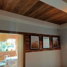 Usług remotowo-wykończeniowe MAG - Drewniany sufit z widoczną strukturą drewna, białe listwy przypodłogowe, ściana z cegły z trzema obrazami w czarnych ramkach, widok na jasny pokój z wiklinową lampą i roślinami.