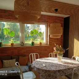 Pokój z ceglaną ścianą, drewnianą zabudową sufitu i pionowymi panelami ściennymi, oświetlony lampami z wikliny, z widokiem na ogród przez okno, z roślinami doniczkowymi na parapecie.