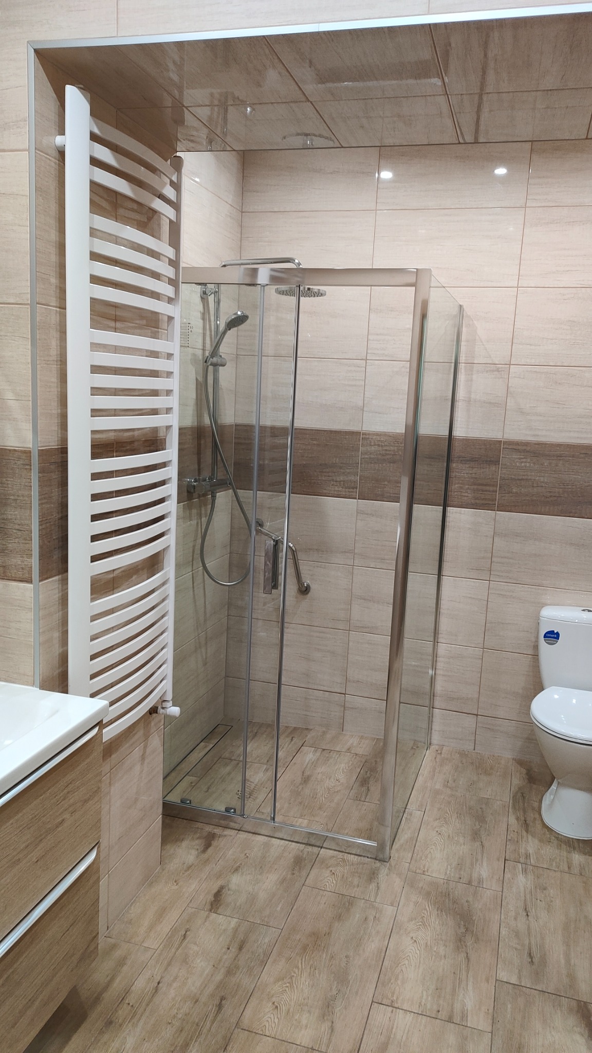 Wykończona łazienka z kabiną prysznicową, grzejnikiem drabinkowym i toaletą, wyłożona płytkami w odcieniach beżu i brązu, z drewnopodobną podłogą.
