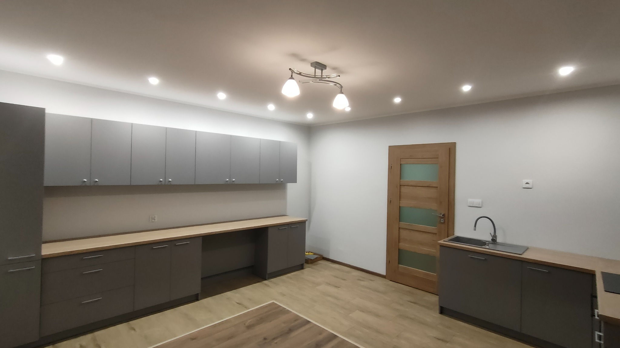 Szare szafki kuchenne z drewnianym blatem, widok na drewniane drzwi z szybami i fragment podłogi z panelami.