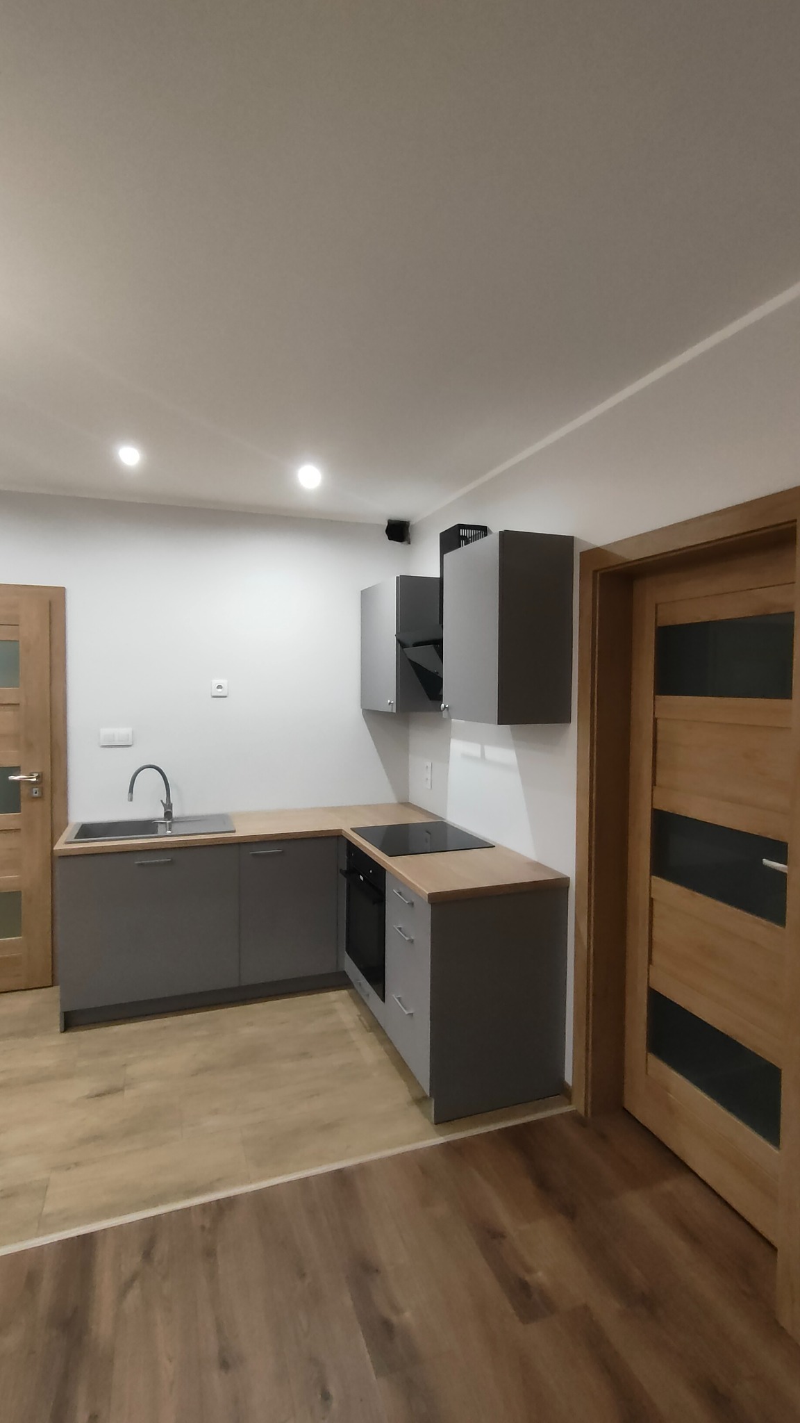Nowoczesna, szara zabudowa kuchenna z drewnianym blatem i płytą indukcyjną, widok na drewnianą podłogę i drzwi z przeszkleniem, oświetlenie punktowe sufitowe.