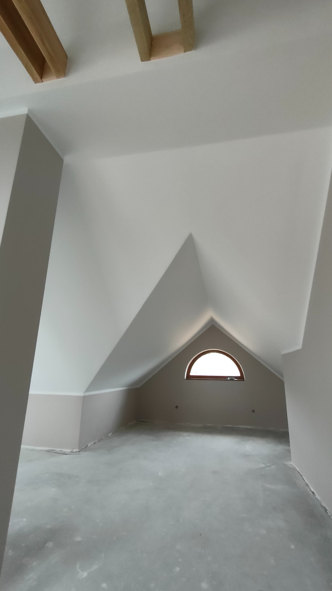 Poddasze po remoncie, świeżo pomalowane ściany w odcieniach szarości, okrągłe okno dachowe, surowa betonowa podłoga, widoczne drewniane elementy konstrukcyjne dachu.