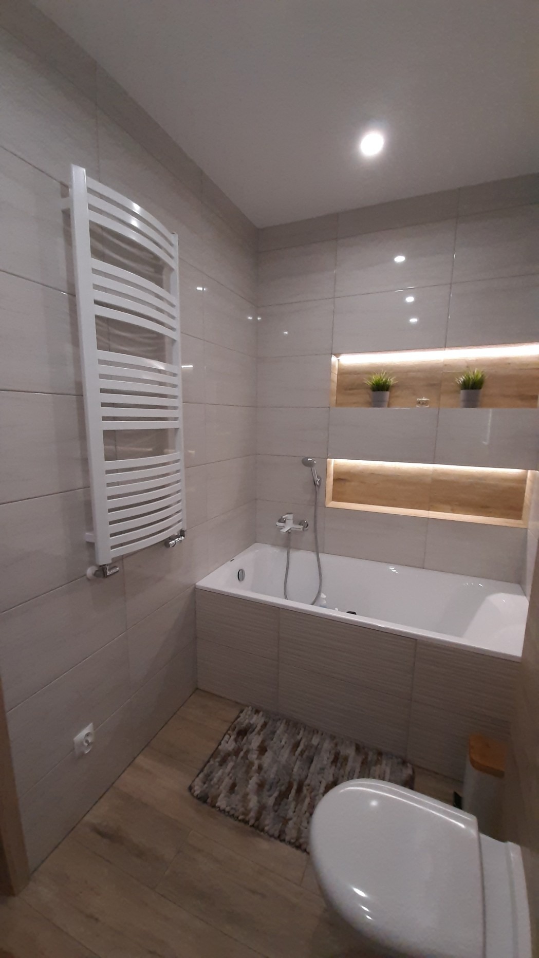 Wykończona łazienka z beżowymi płytkami, białą wanną z obudową z poziomych paneli, grzejnikiem drabinkowym, podświetlanymi wnękami z dekoracjami i dywanikiem na podłodze.