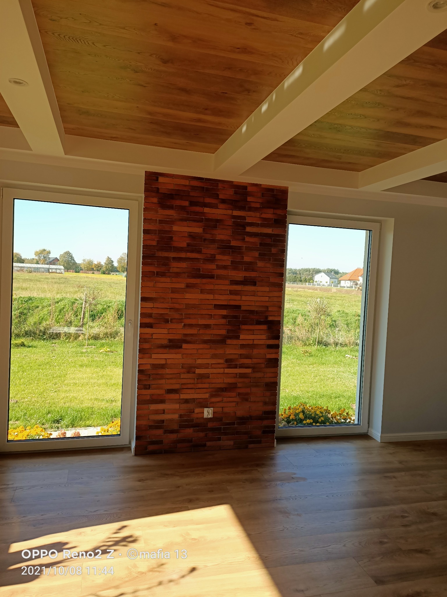 Wykończone wnętrze z drewnianym sufitem kasetonowym, ścianą z cegły klinkierowej pomiędzy dwoma oknami z widokiem na zielony krajobraz i drewnianą podłogą w jodełkę.
