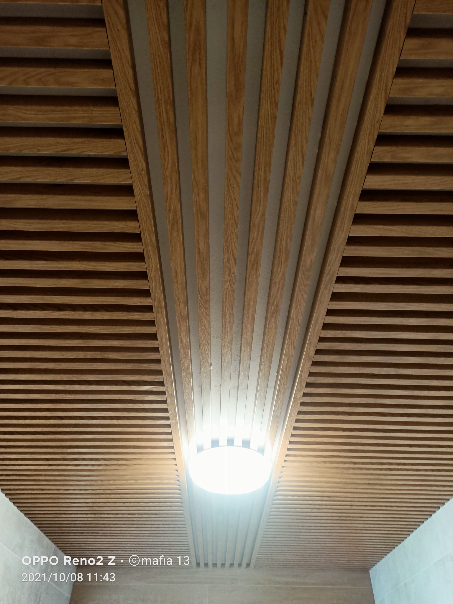 Drewniany sufit z listew ułożonych równolegle, z fragmentem ułożonym pod kątem, podświetlony okrągłą lampą.