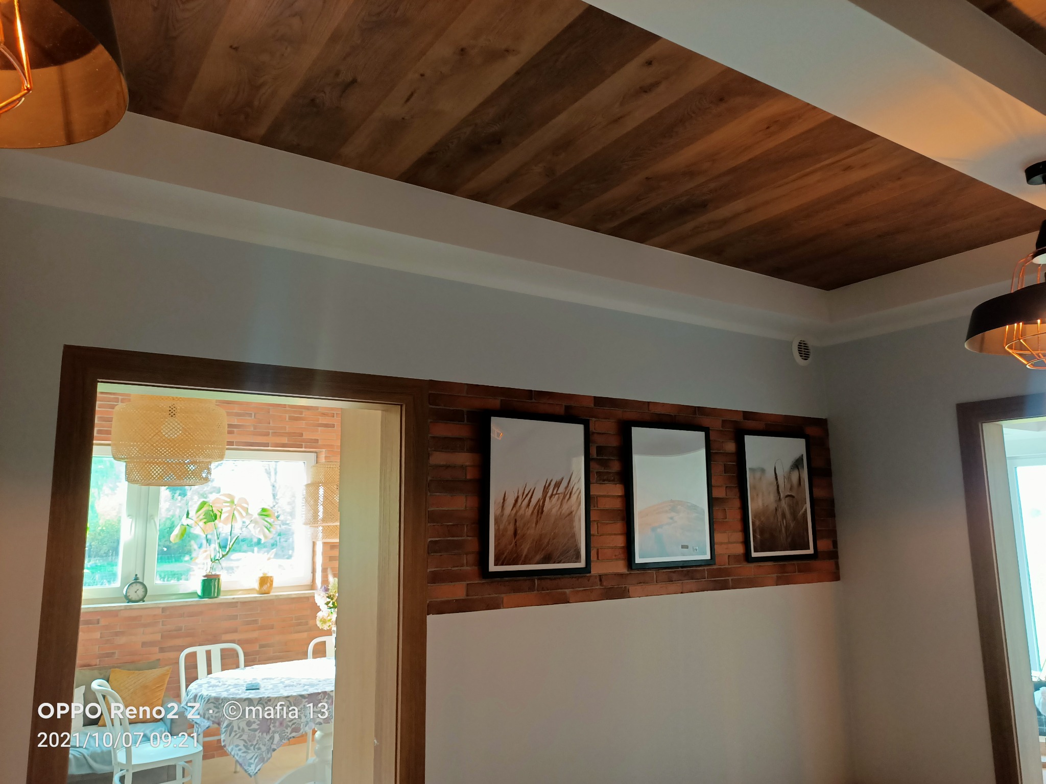 Drewniany sufit z widoczną strukturą drewna, białe listwy przypodłogowe, ściana z cegły z trzema obrazami w czarnych ramkach, widok na jasny pokój z wiklinową lampą i roślinami.