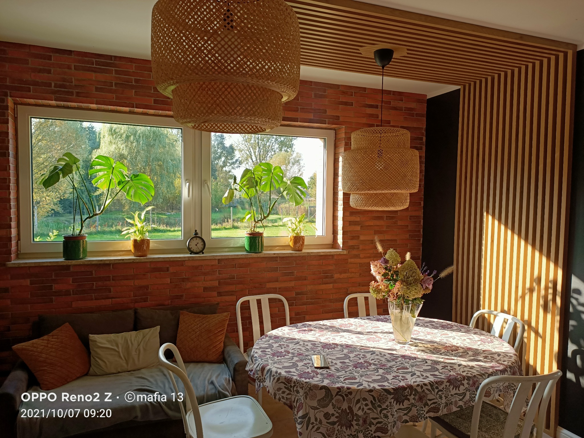 Pokój z ceglaną ścianą, drewnianą zabudową sufitu i pionowymi panelami ściennymi, oświetlony lampami z wikliny, z widokiem na ogród przez okno, z roślinami doniczkowymi na parapecie.