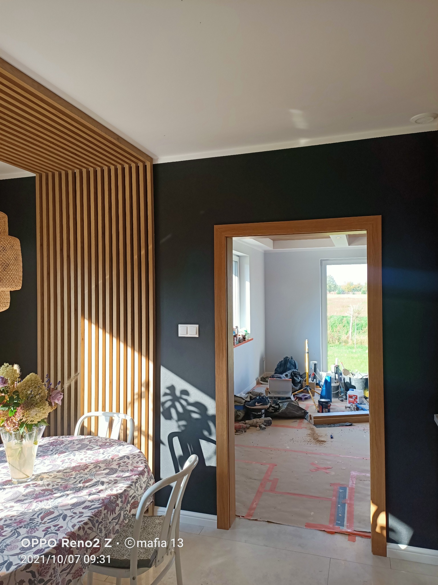 Częściowo wykończone wnętrze z czarną ścianą, drewnianą ażurową ścianką działową i widocznym bałaganem remontowym w kolejnym pomieszczeniu, widoczne narzędzia i materiały budowlane.