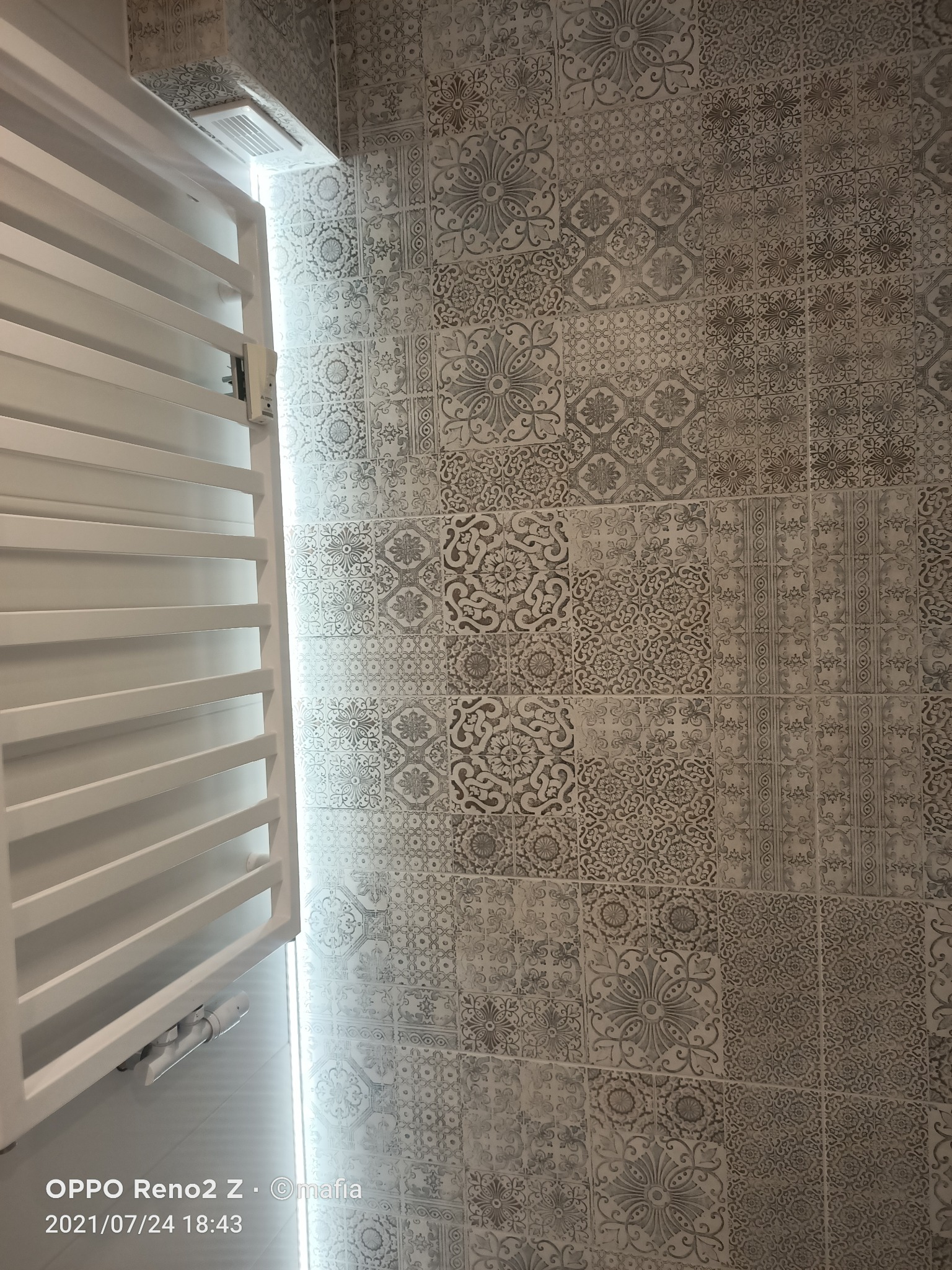 Grzejnik drabinkowy łazienkowy, biały, zamontowany na ścianie wyłożonej dekoracyjnymi, wzorzystymi płytkami ceramicznymi w odcieniach szarości.