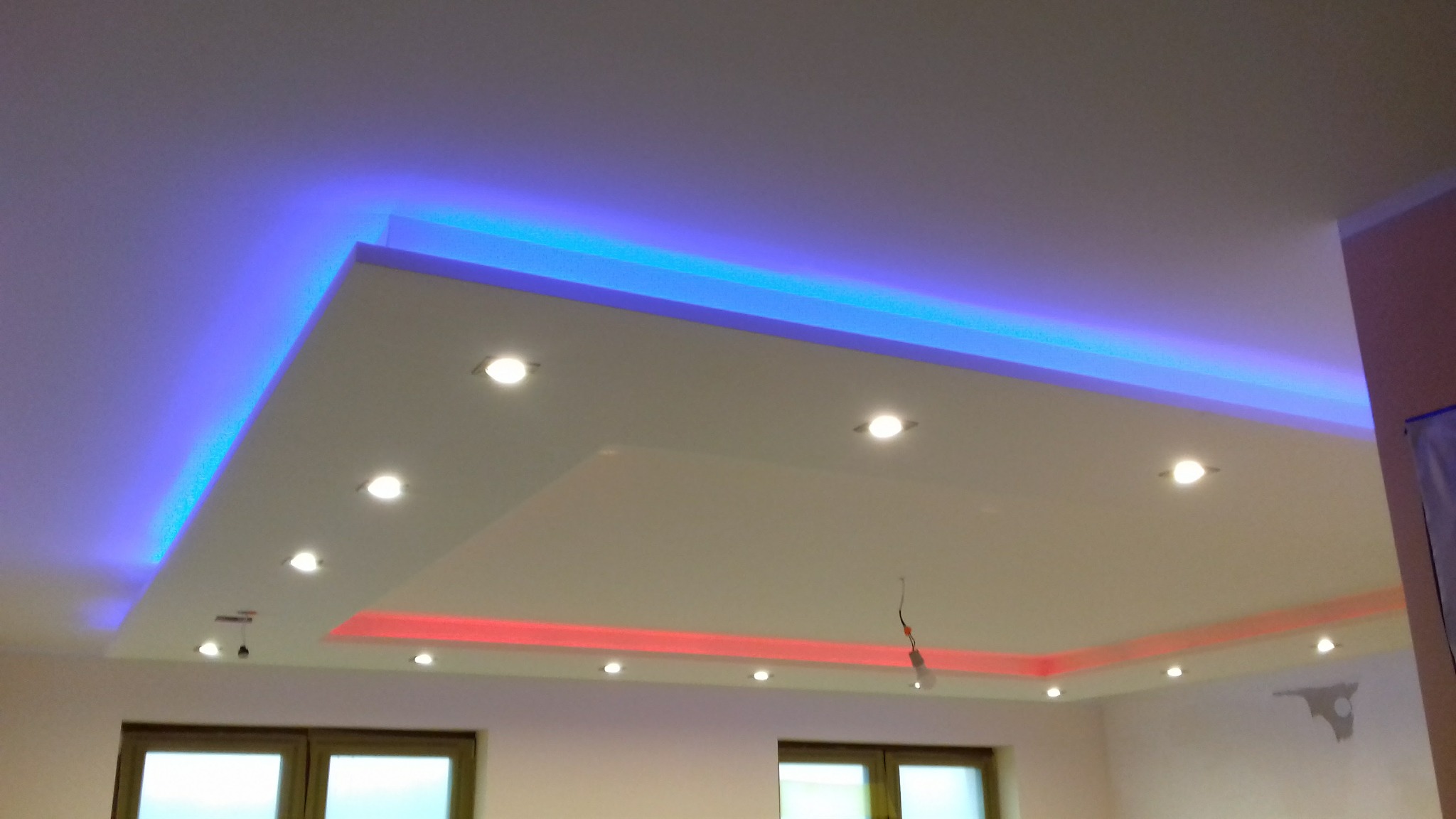 Podwieszany sufit z oświetleniem LED w kolorach niebieskim i czerwonym oraz halogenami, widoczne okna w tle.