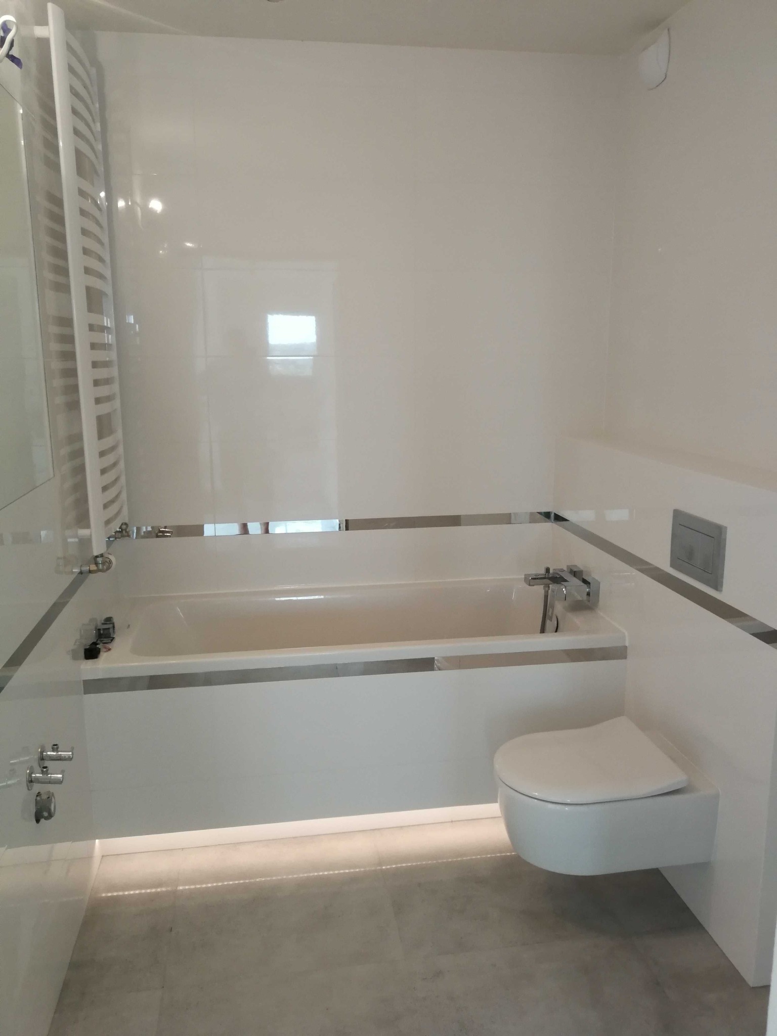 Biała łazienka z wanną obudowaną płytkami, podwieszaną toaletą i grzejnikiem drabinkowym, wykończona srebrnymi listwami dekoracyjnymi, oświetlona taśmą LED pod wanną.