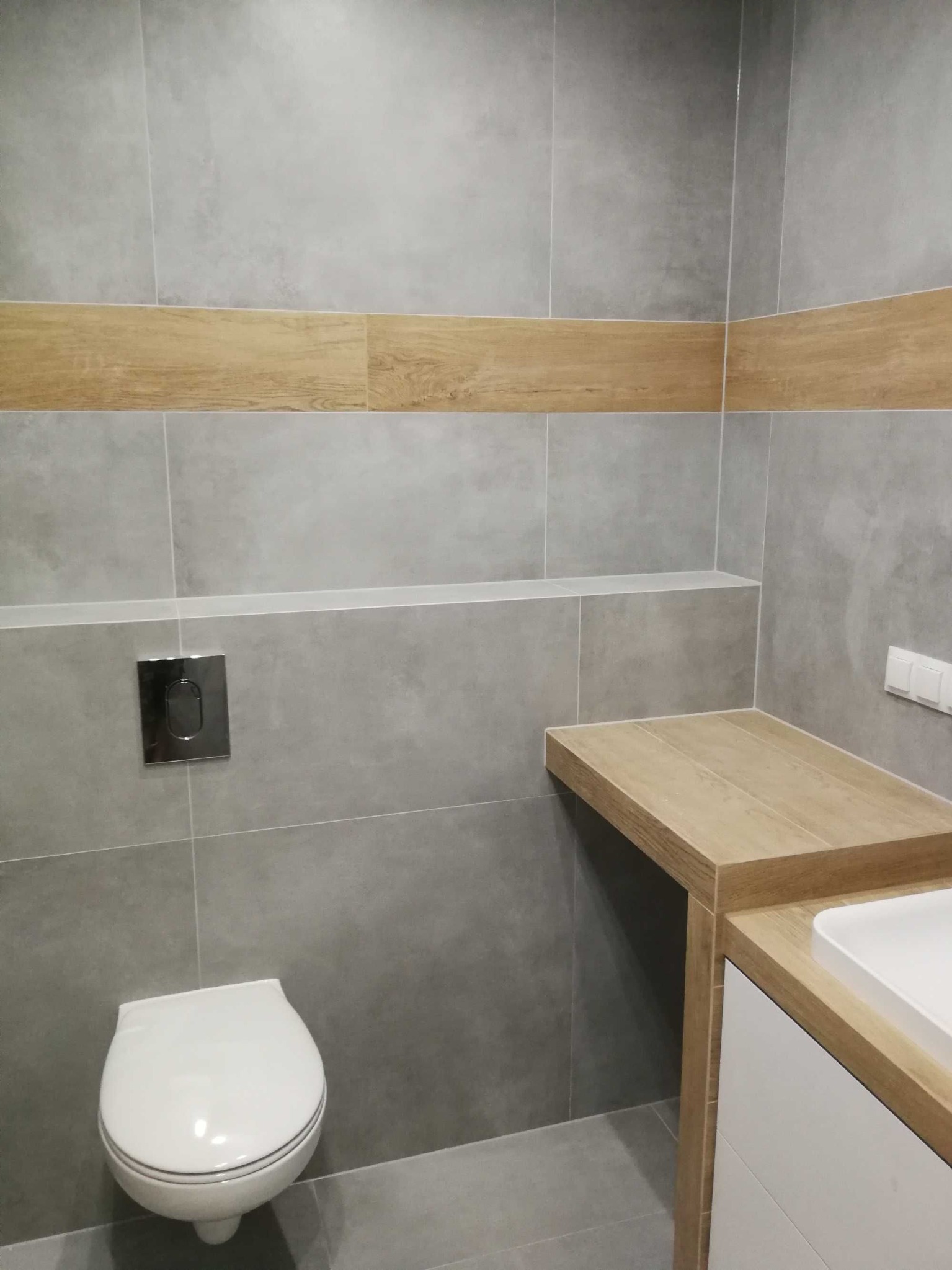 Szara łazienka z drewnopodobnymi akcentami: płytki ścienne, półka, zabudowa umywalki i pas dekoracyjny. Wisząca toaleta z chromowanym przyciskiem spłukiwania. Minimalistyczne oświetlenie.