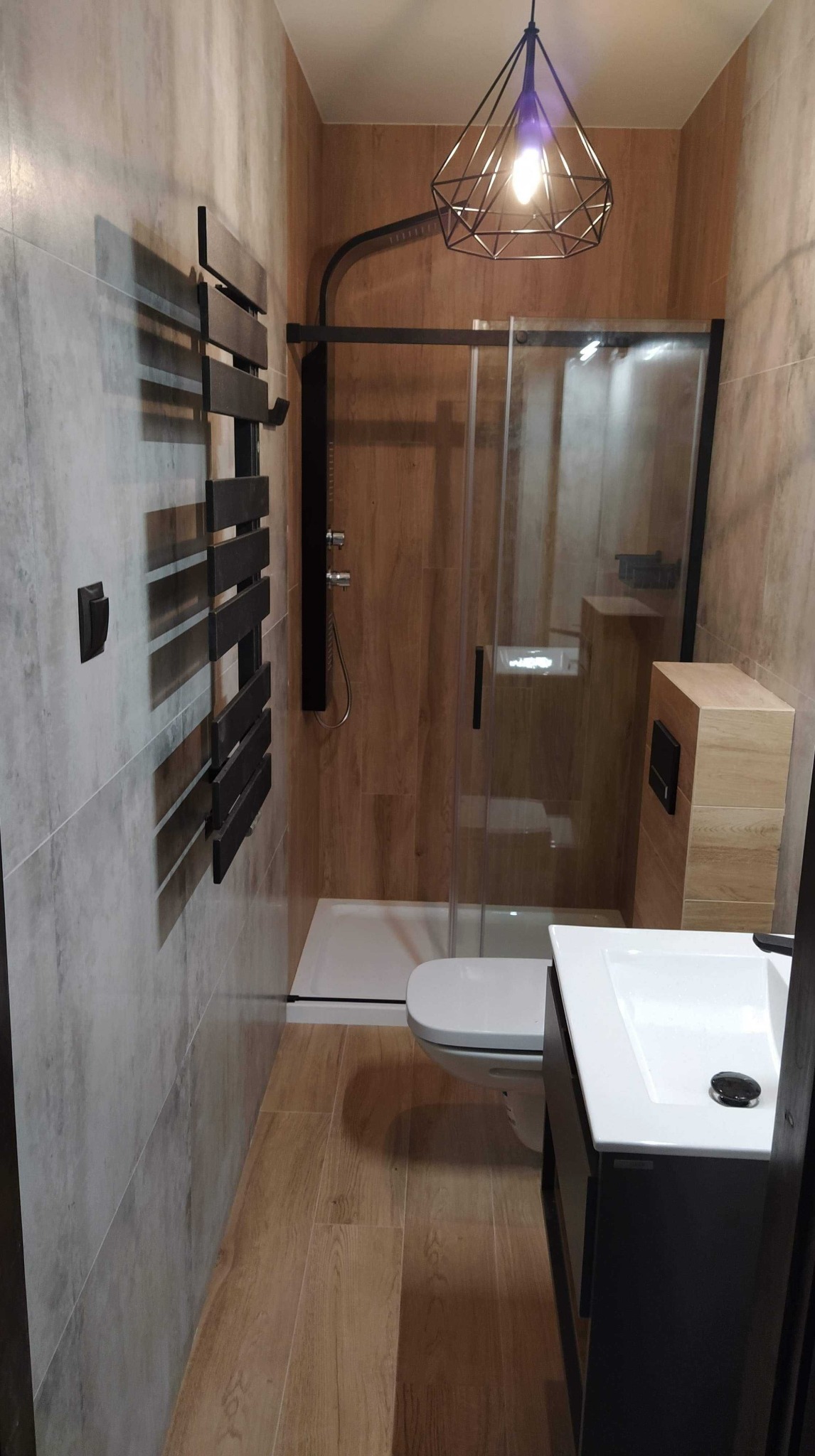 Nowoczesna łazienka z kabiną prysznicową typu walk-in, grzejnikiem drabinkowym w kolorze czarnym, podwieszaną toaletą i umywalką z szafką, oświetlona lampą wiszącą w geometrycznej oprawie. Ściany...