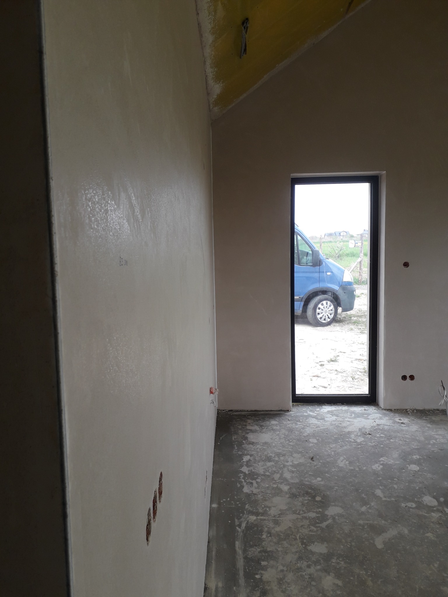 Świeżo nałożony, gładki tynk maszynowy na ścianie wewnętrznej, widoczne puszki elektryczne, przez okno widać zaparkowany niebieski bus.