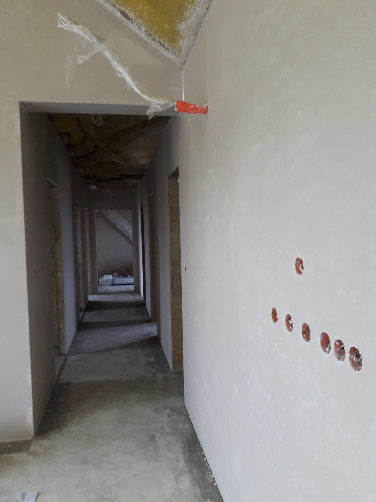 Korytarz w trakcie prac wykończeniowych, ściany pokryte świeżym tynkiem, widoczne otwory pod instalacje elektryczne, folia ochronna na suficie.