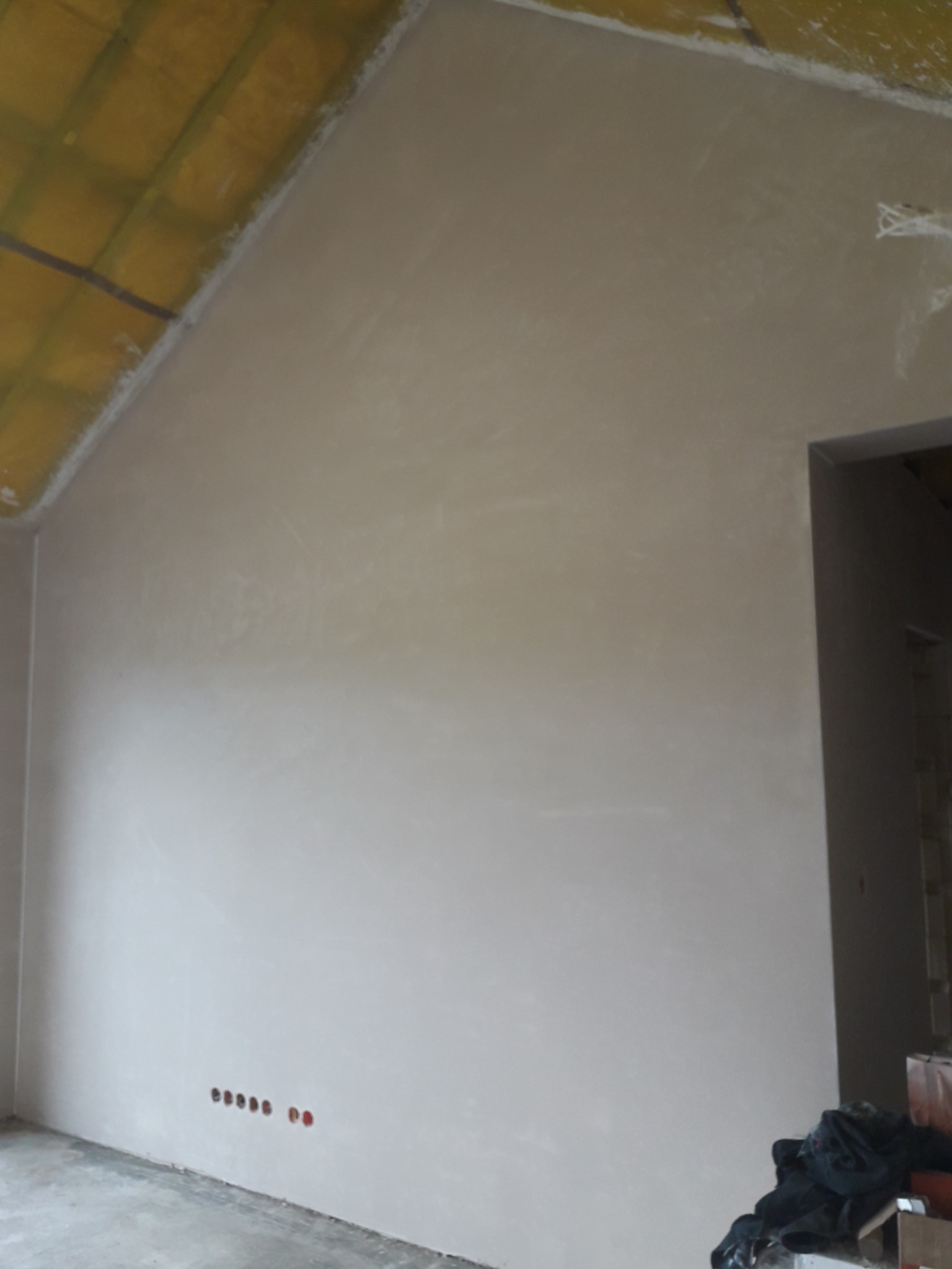 Świeżo nałożona, gładka, jasna warstwa tynku na ścianie poddasza z widocznymi otworami na gniazdka elektryczne i żółtą izolacją dachu.