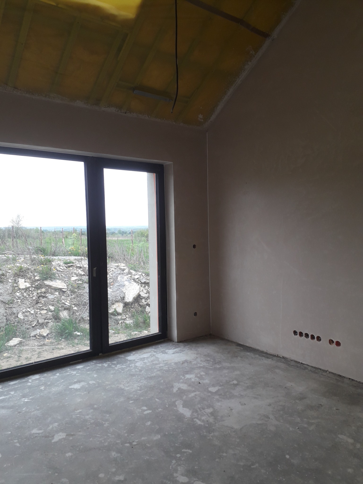 Wnętrze surowego domu po nałożeniu gładzi, widoczne puszki elektryczne, okno z widokiem na zewnątrz i żółta folia na poddaszu.