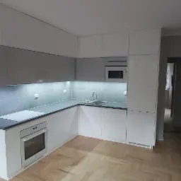 Nowoczesna, narożna kuchnia w odcieniach szarości i bieli z drewnianą podłogą w jodełkę, oświetleniem podszafkowym LED i wbudowanym piekarnikiem.