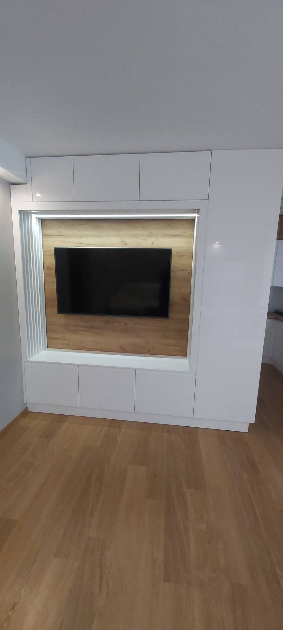 Biała zabudowa meblowa z miejscem na telewizor, wykończona drewnopodobną płytą i oświetleniem LED, widoczna na tle drewnianej podłogi.