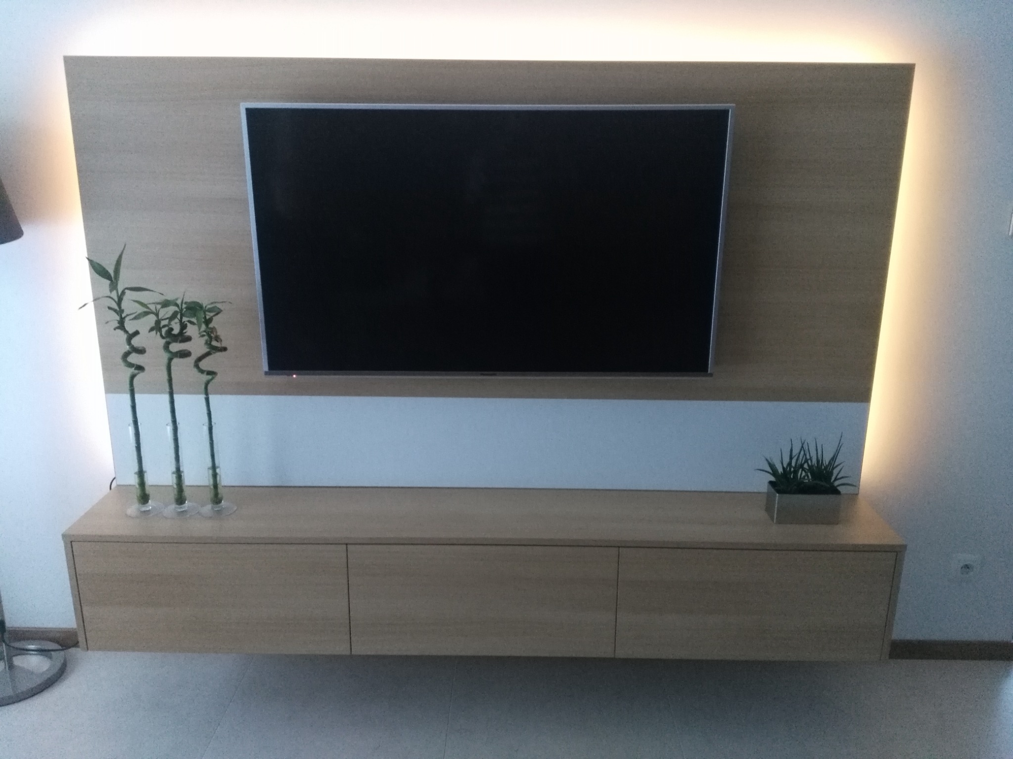 Drewniana szafka RTV z trzema frontami, zamontowana pod telewizorem na ścianie z podświetleniem LED, dekorowana bambusami w szklanych naczyniach i sukulentami w metalowej doniczce.