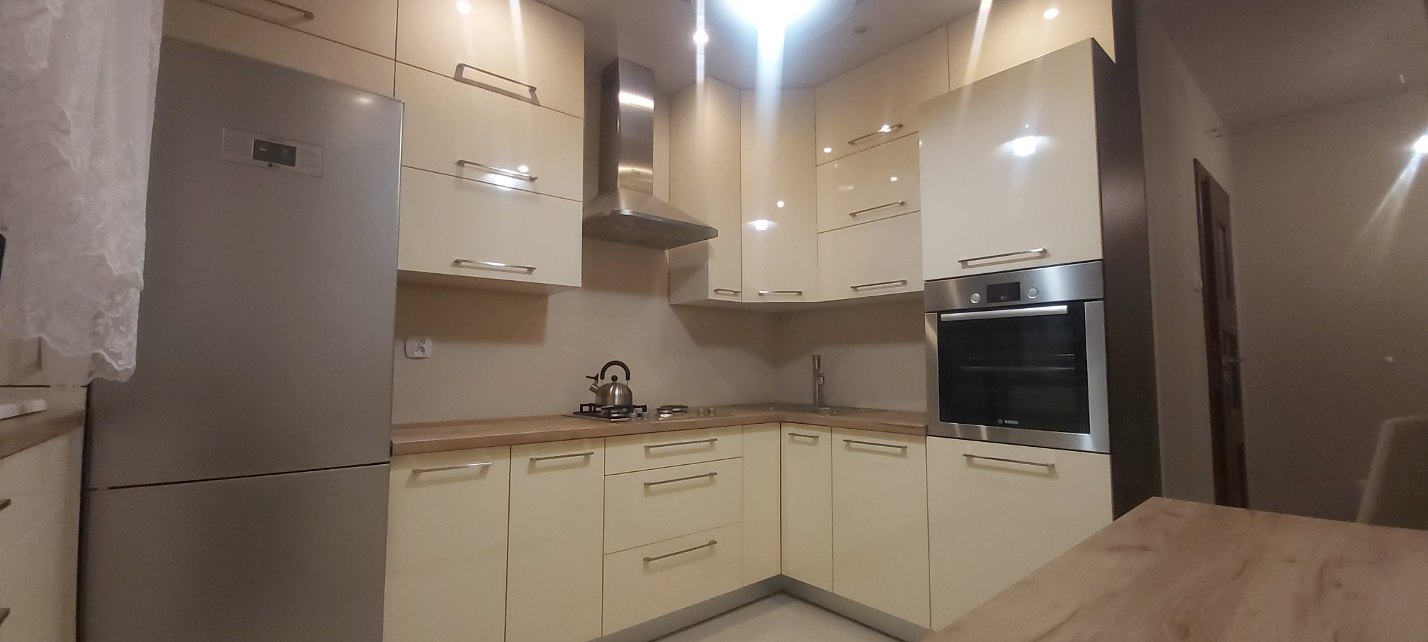 Jasna kuchnia z połyskującymi frontami, srebrną lodówką i okapem, oraz wbudowanym piekarnikiem. Na blacie czajnik na płycie gazowej.