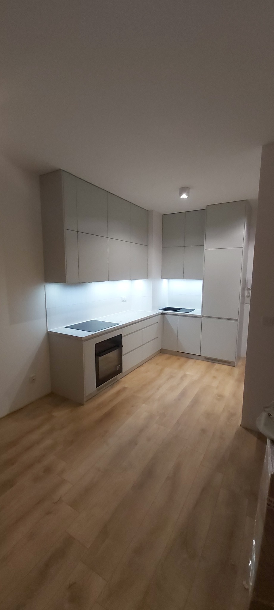 Jasnoszara, minimalistyczna zabudowa kuchenna z białymi blatami i drewnianą podłogą, oświetlona od dołu, widok z rogu pomieszczenia.
