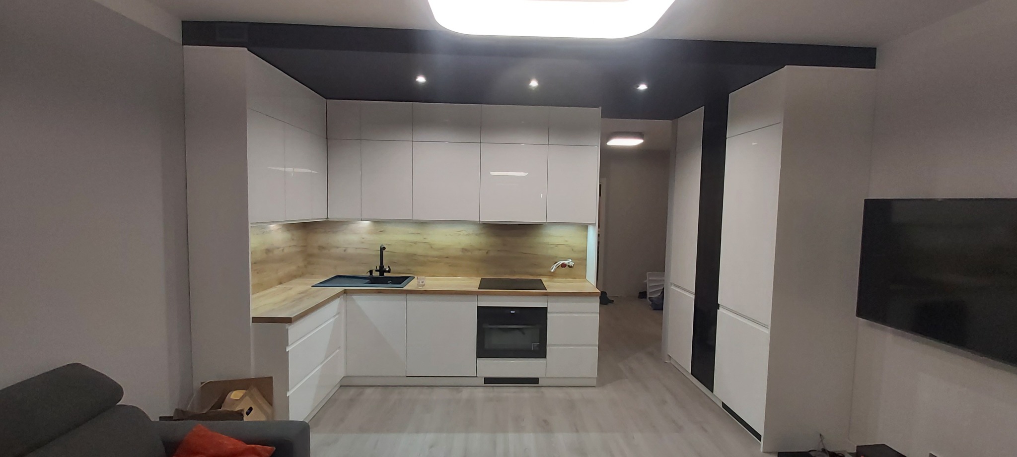 Nowoczesna kuchnia z białymi lakierowanymi frontami, drewnianym blatem i czarną armaturą, oświetlona punktowymi lampami pod szafkami i centralnym plafonem.