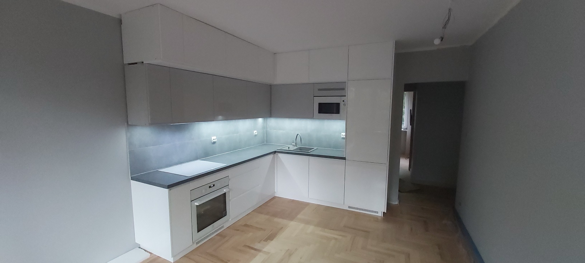 Nowoczesna, narożna kuchnia w odcieniach szarości i bieli z drewnianą podłogą w jodełkę, oświetleniem podszafkowym LED i wbudowanym piekarnikiem.