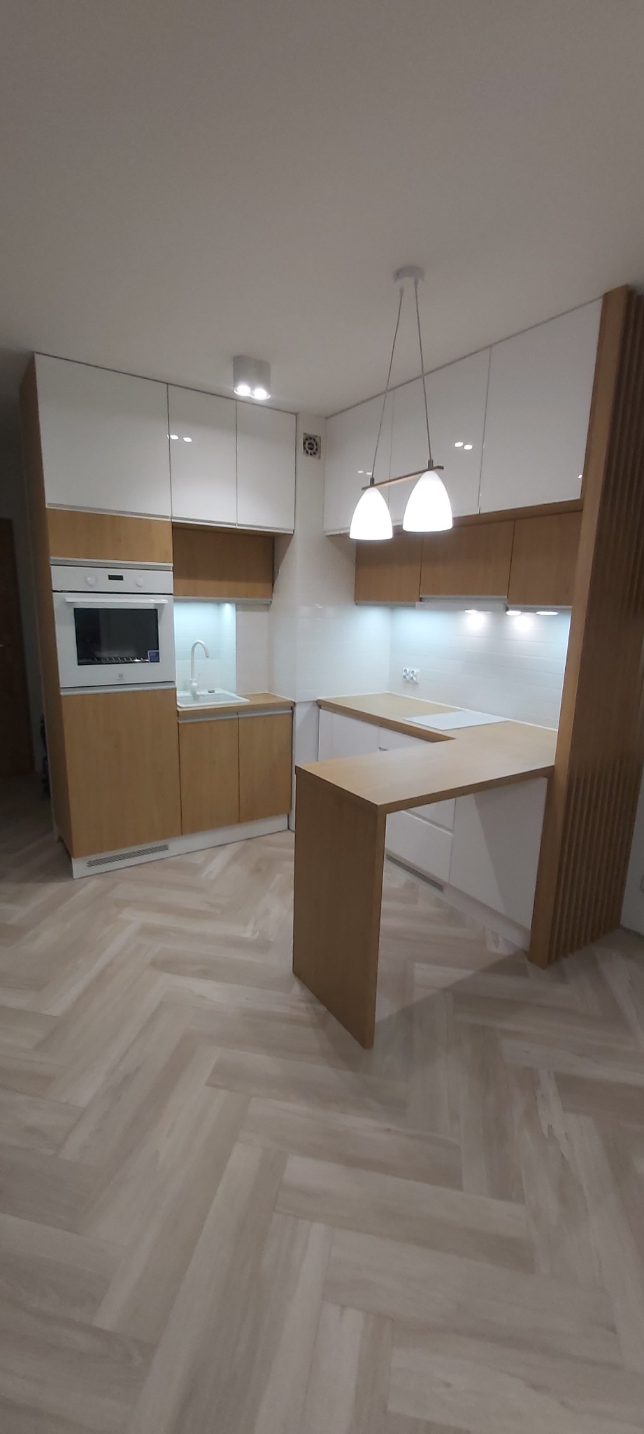 Nowoczesna kuchnia z białymi, błyszczącymi szafkami górnymi i drewnianymi szafkami dolnymi, oświetlona wiszącymi lampami, z blatem przechodzącym w barek, na podłodze parkiet ułożony w jodełkę.