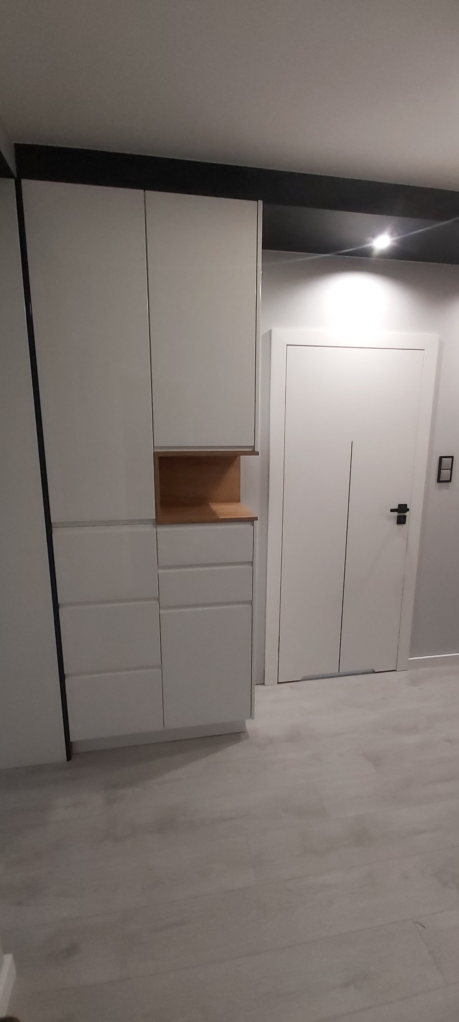 Biała szafa na wymiar z drewnianą wnęką, obok białe drzwi z czarną klamką i listwą przypodłogową, na jasnej podłodze.