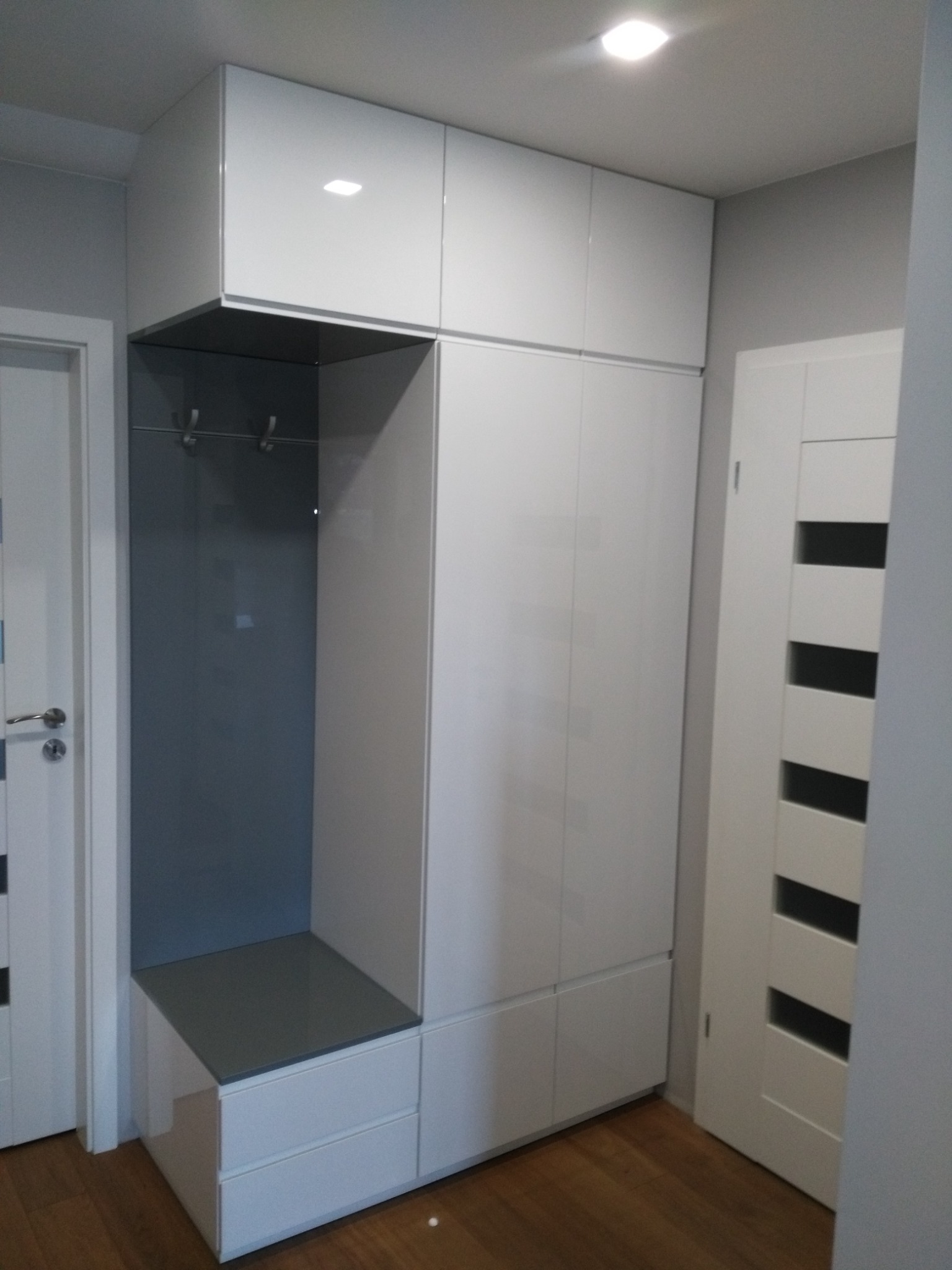 Biała szafa w przedpokoju z siedziskiem i wieszakami, fronty lakierowane na wysoki połysk, drewniana podłoga.